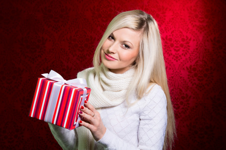 Portrait of casual young happy smiling blonde hold striped gift box. Isolated studio red background female model.の写真素材