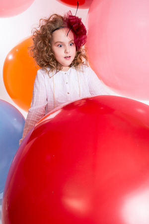 Young teen girl in a hat and white dress on a background of big colored rubber air balls. Cheerful girl looks at the big red ballの写真素材