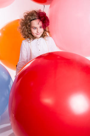 Young teen girl in a hat and white dress on a background of big colored rubber air balls. Surprised girl looking at a big red ballの写真素材