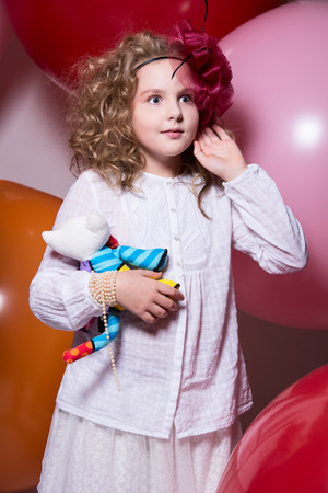 Surprised girl in a red hat in a white dress with a soft toy in his hand looking to the side. Against the background of large rubber balloonsの写真素材