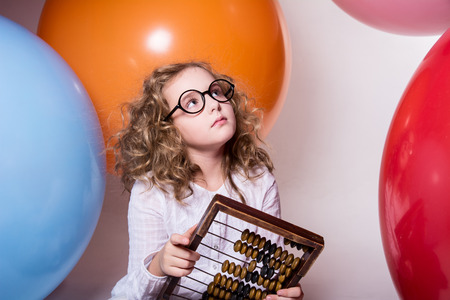 Thoughtful curly teen girl in glasses with wooden abacus on the background of large rubber balls. Girl looking upの写真素材