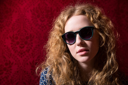 Close-up portrait of beautiful pensive girl with curly hair wearing sunglasses. A woman on a dark red の写真素材