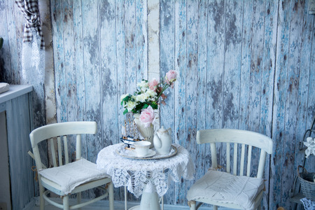 The interior in blue tones with two chairs and a vase with flowers. On the table are a kettle and a cup of tea. Wall of old boardsの写真素材