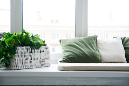 Pillows on the windowsill and plastic window. Wicker basket with flowers on the windowsill. View from the window on the house across the streetの写真素材