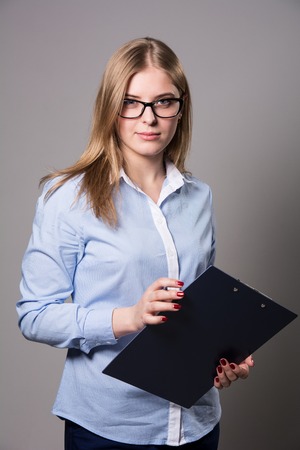 Pensive young woman in glasses standing and holding clipboard and pen over grau backgroundの写真素材