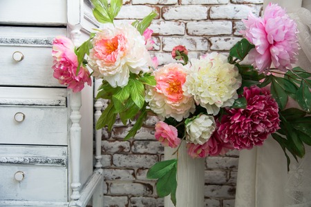 Artificial flowers in a vase standing next to an old chest of drawers against the wall