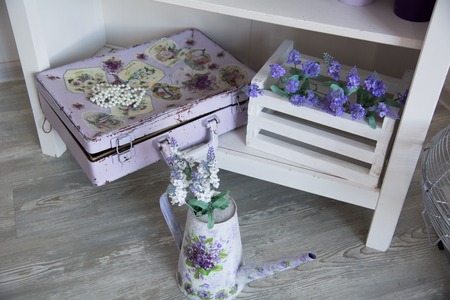 Painted suitcase and a watering can with flowers on the floor. Lilac flowers in a box on a shelf in a country houseの写真素材