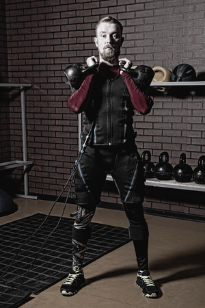 Red-haired man in an electric muscular suit for stimulation with weights. Fitness in the EMC suit in the training room against the backdrop of a brick wallの写真素材