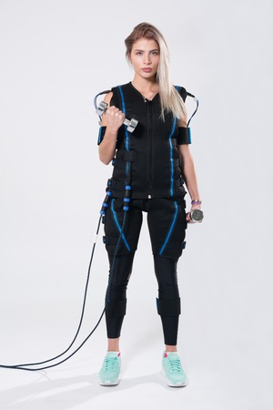 Beautiful blonde in electric muscular suit for stimulation with dumbbells. Young woman in EMC suit isolated on white backgroundの写真素材