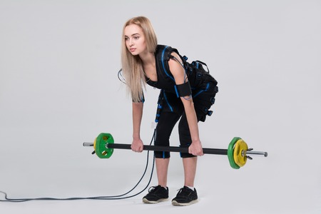Beautiful blonde in an electric muscular suit for stimulating exercises with a barbell. Young woman in EMC suit isolated on white backgroundの写真素材