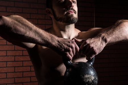 Close-up of a muscular man lifts kettlebell. Fitness man with a beard against a brick wall...