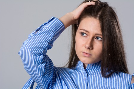 Pensive young woman holding her head looking away. Girl with long hair and striped shirt on a gray backgroundの写真素材