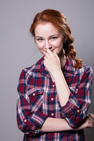 Young red-haired girl in a plaid shirt with fingers on his lip. Beautiful fashion woman smiling isolated in studio on a gray background.の写真素材
