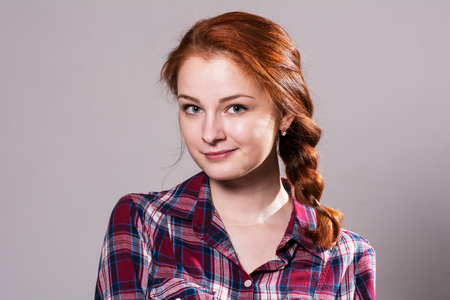 Close-up portrait of a girl looking at camera. Red-haired girl with a pigtail smiling in a plaid shirt on a gray backgroundの写真素材