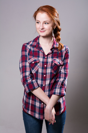 Portrait of a smiling young woman in a plaid shirt. A cute red-haired girl with her arms in front on a gray backgroundの写真素材
