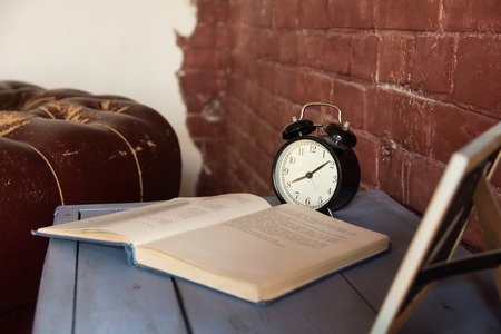 Desk clock with an open book on a wooden table against a brick wall background.
