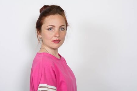Portrait of a red-haired beautiful serious woman looking into the camera on a white background. Positive girl in pink topの写真素材