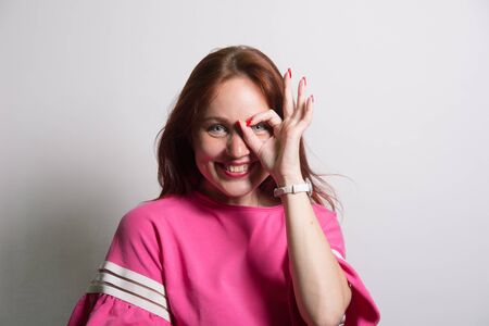 Close up portrait of beautiful joyful redhead Caucasian female smiling, demonstrating white teeth, looking at the camera through fingers in okay gesture. Face expressions, emotions, and body languageの写真素材