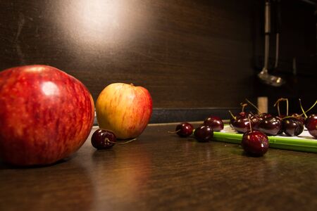 Background of big apples and dark cherries on the kitchen table. Fruit in the kitchen and an illuminated brown wallの写真素材