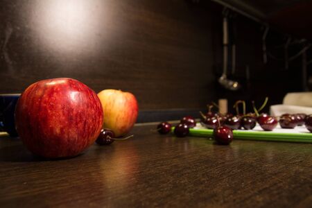 Background apples and dark cherries on the kitchen table. Fruit in the kitchen and an illuminated brown wallの写真素材