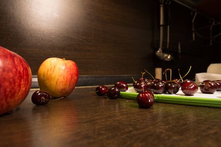 Background apples and dark cherries on the kitchen table. Fruit in the kitchen and an illuminated brown wallの写真素材