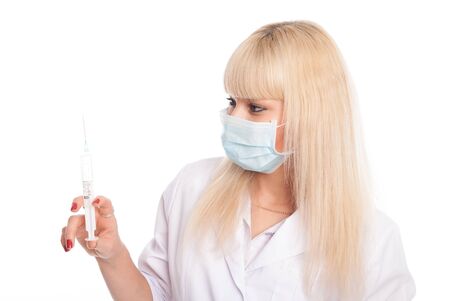 Close-up of a young blonde nurse in a mask looking at a syringe. Doctor epidemic isolated over a white backgroundの写真素材