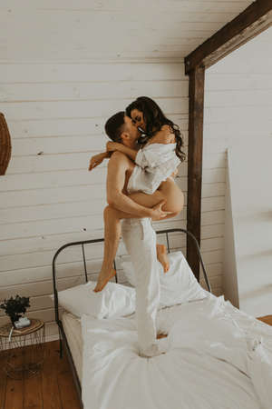 Beautiful young couple in love on the bed in the bedroom. The guy holds the girl in his arms. Kiss each other.の写真素材