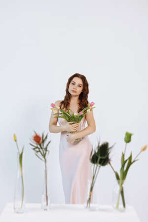 A young girl in a pale pink dress sits on a white cube in the studio. Around her are fresh flowers in vases.の写真素材