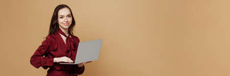 Cheerful young woman standing isolated on beige background and using laptop.の写真素材