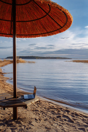 An open laptop lies on a bench under a straw umbrella.の写真素材