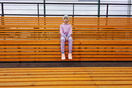 A girl sits on a bench in a sports tribune with her hands on her knees.