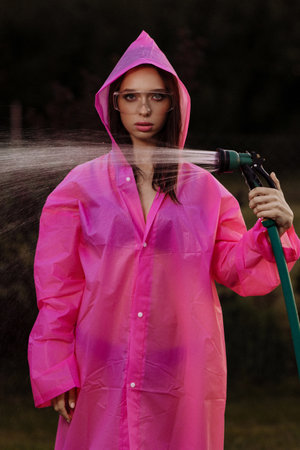 A glamour woman in a pink raincoat is spraying water from a hoseの写真素材