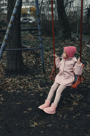 Sad little girl on a pink shabby swing in a bad neighborhood.の写真素材