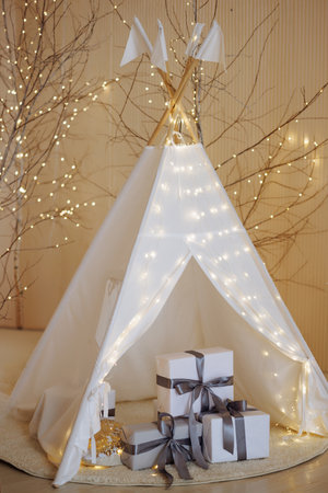 Christmas interior. White wigwam illuminated with lights, and gifts are stacked inside.