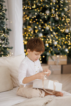 A young boy sits on a bed with a white blanket and a white shirt, opening a present.の写真素材