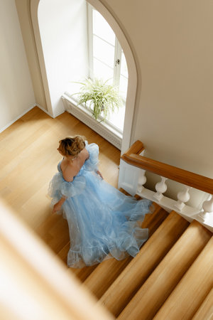 Young woman in light blue evening dress climbs large wooden staircase in luxurious manor. Top view.の写真素材