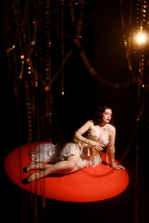 Woman in vintage corset lying on red stage floor under hanging beads on dark backgroundの写真素材