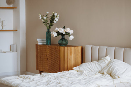 Bright bedroom with unmade white bed, wooden cabinet and flowers in glass vases on beige wallの写真素材