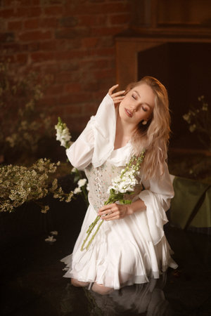 Young woman in white dress sitting in artificial pond with flowers and fog against brick wallの写真素材
