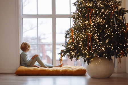 Girl sitting on cushion by window near decorated Christmas tree with lights in cozy roomの写真素材