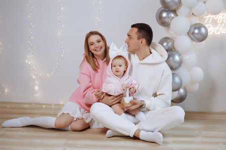 Happy young family with baby celebrating birthday on wooden floor near white wall and balloons