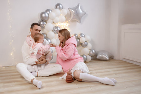 Happy young family with baby celebrating birthday on wooden floor near white wall and balloons