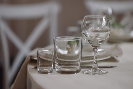 Empty glassware on elegant table with beige tablecloth and white chairs in soft lightの写真素材