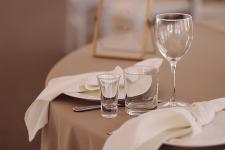 Empty glassware on elegant table with beige tablecloth and white chairs in soft lightの写真素材