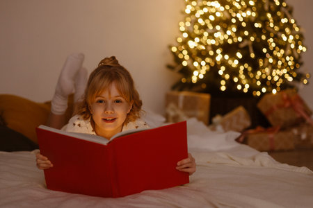 Little girl lying on bed reading red book near Christmas tree with lights and giftsの写真素材