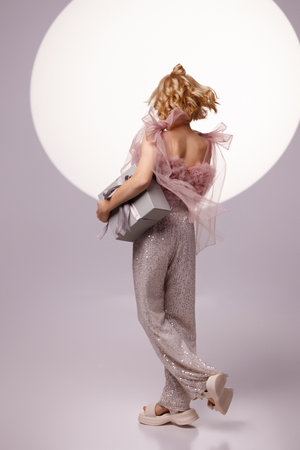 Girl holding Christmas gift box in shiny outfit on light background rear viewの写真素材