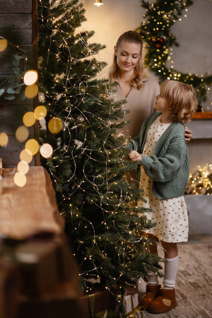 Caucasian mother and daughter decorating Christmas tree in cozy living room with warm lightsの写真素材