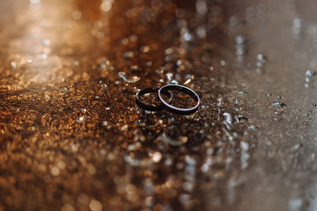 Two wedding rings on wet reflective surface with water droplets and golden bokeh lights
