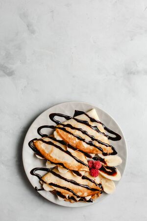 Pancakes with banana, chocolate and raspberries on a plate on a light concrete background. The view...