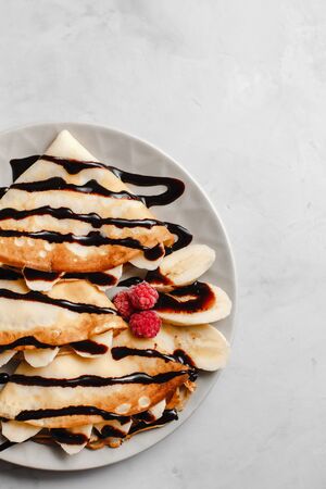 Pancakes with banana, chocolate and raspberries on a plate on a light concrete background. The view from the top. side view. space for textの写真素材